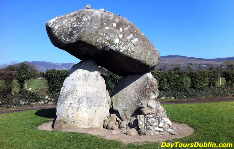 Proleek Dolmen portal tomb with a massive rounded capstone on upright stones in a grassy field, hills behind a hedge under a blue sky.